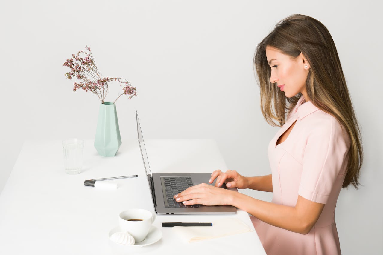our-story-01 A woman in a pink dress working on a laptop at a modern, minimalist desk.