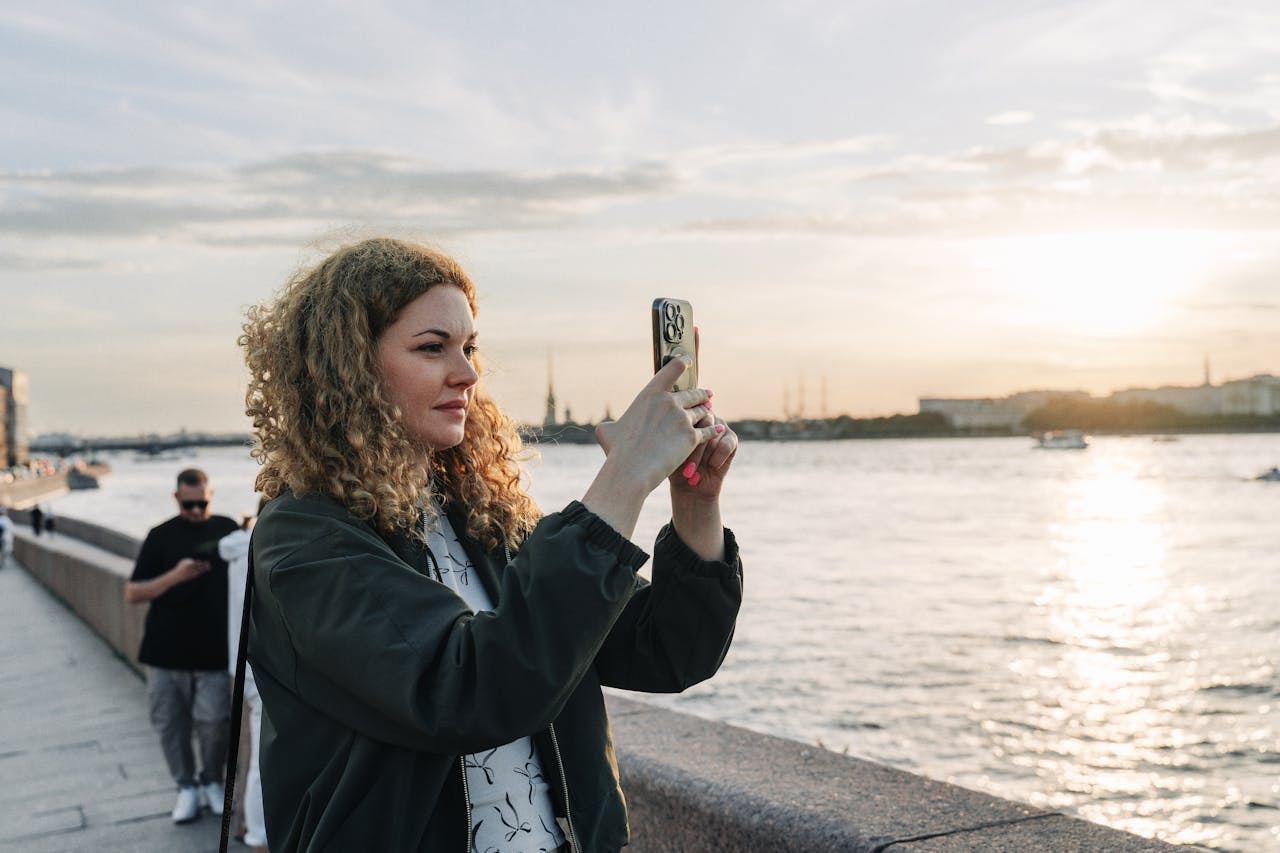 about-01 A woman captures sunset on her smartphone by the waterfront with scenic views.